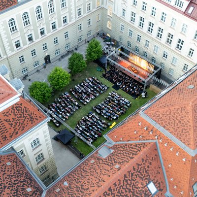 Zuschauerinnen sitzen bei einer Open‑Air‑Veranstaltung und blättern in Programmbroschüren; eine Person hält ein Getränk.