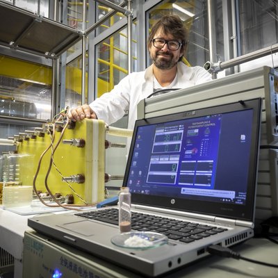 Ein Mann steht in einem Labor hinter einem Stromspeicher und einem Laptop.