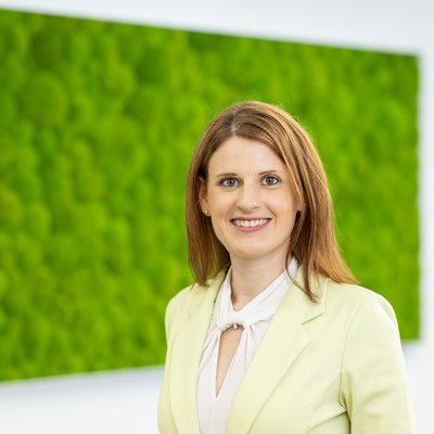      Porträt einer lächelnden Frau im hellgrünen Blazer vor einer großen grünen Mooswand in einem hellen Innenraum.