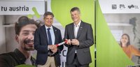 Zwei Herren in Anzügen lächeln in die Kamera. Der linke übergibt dem rechten ein Staffelholz.