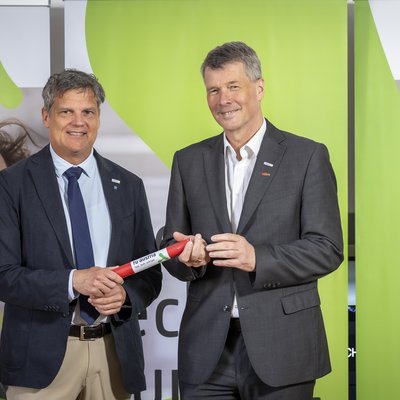 Zwei Herren in Anzügen lächeln in die Kamera. Der linke übergibt dem rechten ein Staffelholz.