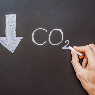 Hand schreibt mit Kreide „CO₂“ auf eine Tafel; daneben ein nach unten zeigender Pfeil – Symbol für sinkende CO₂-Emissionen.