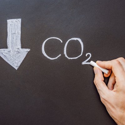 Hand schreibt mit Kreide „CO₂“ auf eine Tafel; daneben ein nach unten zeigender Pfeil – Symbol für sinkende CO₂-Emissionen.