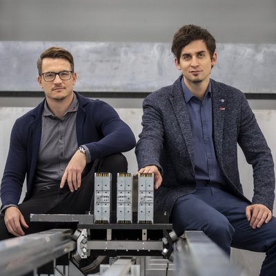 Zwei Männer sitzen auf den Schienen eines Prüfstandes und schauen in die Kamera