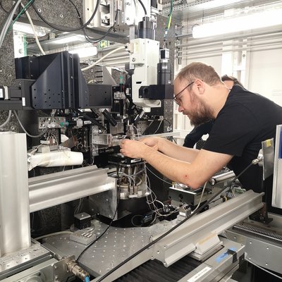 In einem High‑Tech‑Labor arbeitet eine Person konzentriert an einer Experimentierstation mit optischen Modulen, Probenhalter, Motorachsen und zahlreichen Kabeln – typische Ausstattung einer Synchrotron‑Beamline.