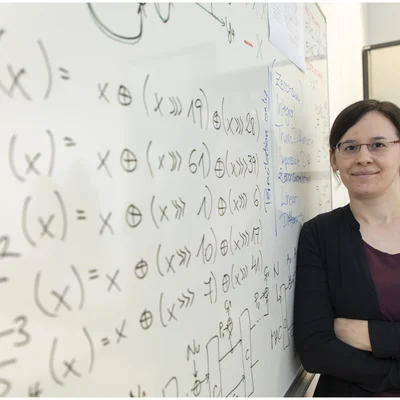 Lächelnde Frau lehnt an Tafel mit mathematischen Symbolen.