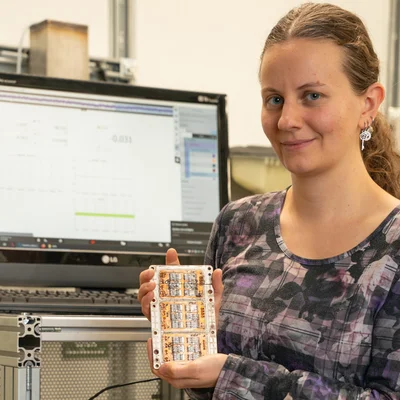 Frau mit elektronischem Steuerelement in der Hand, im Hintergrund ein Computer-Bildschirm. 
