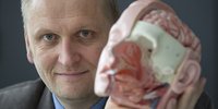 Mann mit graublauen Augen hält die Nachbildung eines halb geöffneten menschlichen Kopfes in die Kamera.