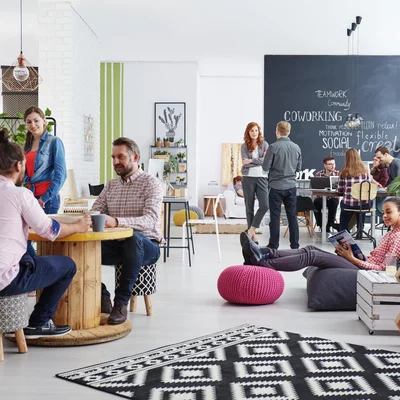 modernes Büro, links im Vordergrund plaudern zwei Männer, schräg dahinter steht eine Frau und hört zu; rechts von der Gruppe sitzt eine junge Frau auf einem bequemen Hocker und liest, im Hintergrund sieht man eine schwarze Tafel auf der mit Kreide Begriffe zum Thema Work stehen, Gruppen von Menschen stehen und sitzen davor