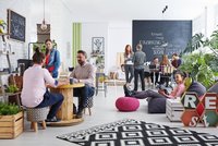 modernes Büro, links im Vordergrund plaudern zwei Männer, schräg dahinter steht eine Frau und hört zu; rechts von der Gruppe sitzt eine junge Frau auf einem bequemen Hocker und liest, im Hintergrund sieht man eine schwarze Tafel auf der mit Kreide Begriffe zum Thema Work stehen, Gruppen von Menschen stehen und sitzen davor