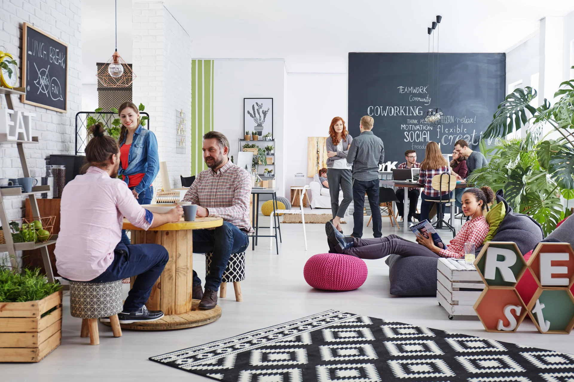 modernes Büro, links im Vordergrund plaudern zwei Männer, schräg dahinter steht eine Frau und hört zu; rechts von der Gruppe sitzt eine junge Frau auf einem bequemen Hocker und liest, im Hintergrund sieht man eine schwarze Tafel auf der mit Kreide Begriffe zum Thema Work stehen, Gruppen von Menschen stehen und sitzen davor