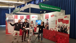 An exhibition stand with the inscription "Graz University of Technology" and large TU Graz advertising spaces. 2 young counsellors and a female counsellor in high spirits at a bar table with information material.