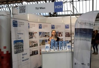 Bereit für Interessierte: Die TUW-Studienassistentin winkend hinter dem Infostand der TU Wien.