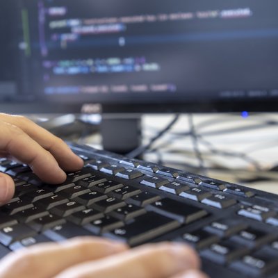 Hände tippen auf einer Computertastatur. Im Hintergrund ist verschwommen ein Computermonitor mit Code darauf zu sehen.