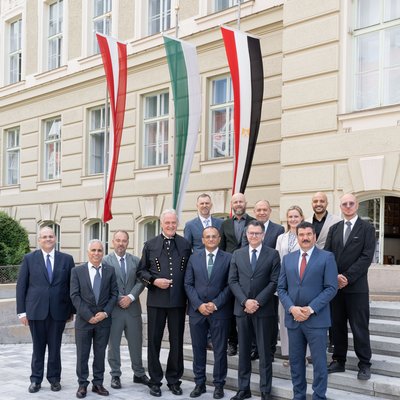 Group photo of representatives from two institutions in front of a historic building with three vertically displayed flags, including those of Austria and Egypt.