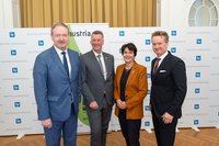 Vier Personen stehen nebeneinander vor einer Logo-Wand.