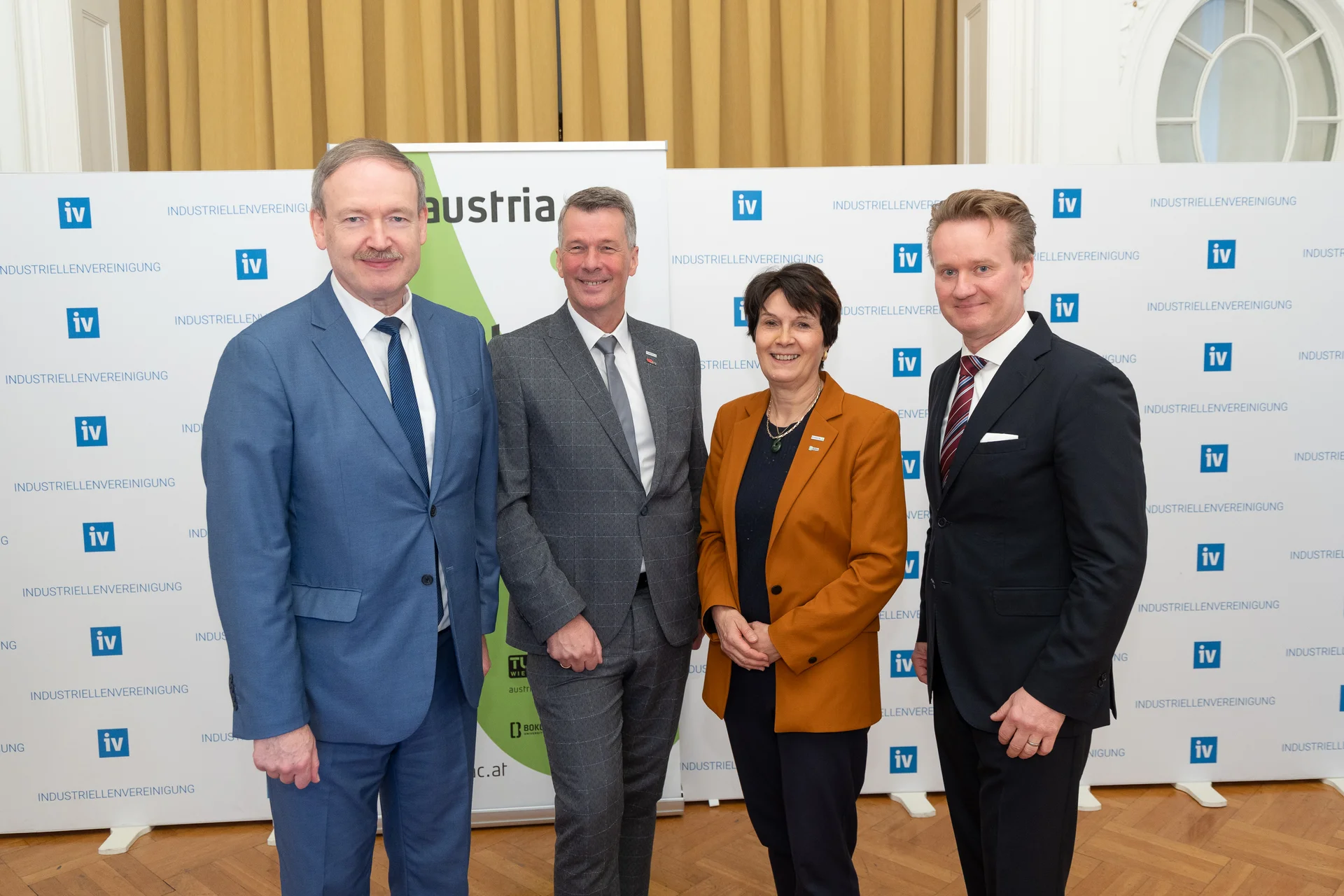 Vier Personen stehen nebeneinander vor einer Logo-Wand.