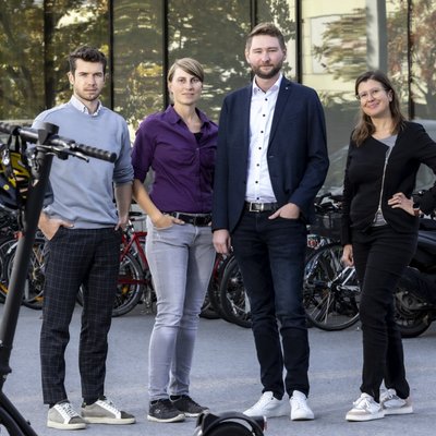 Vier Menschen, zwei Frauen und zwei Männer, schauen in die Kamera. Vor ihnen steht ein E-Scooter