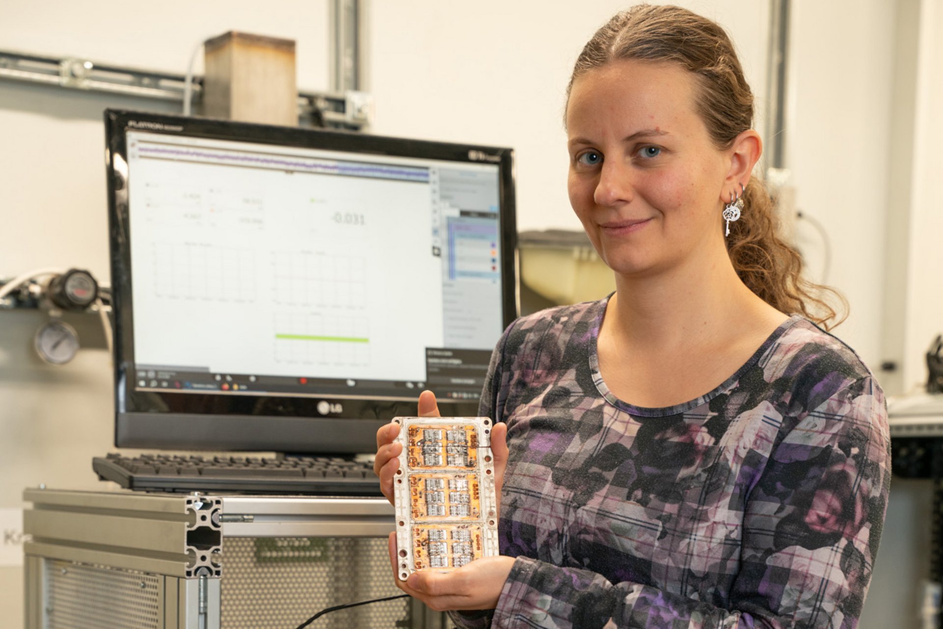 Frau mit elektronischem Steuerelement in der Hand, im Hintergrund ein Computer-Bildschirm.