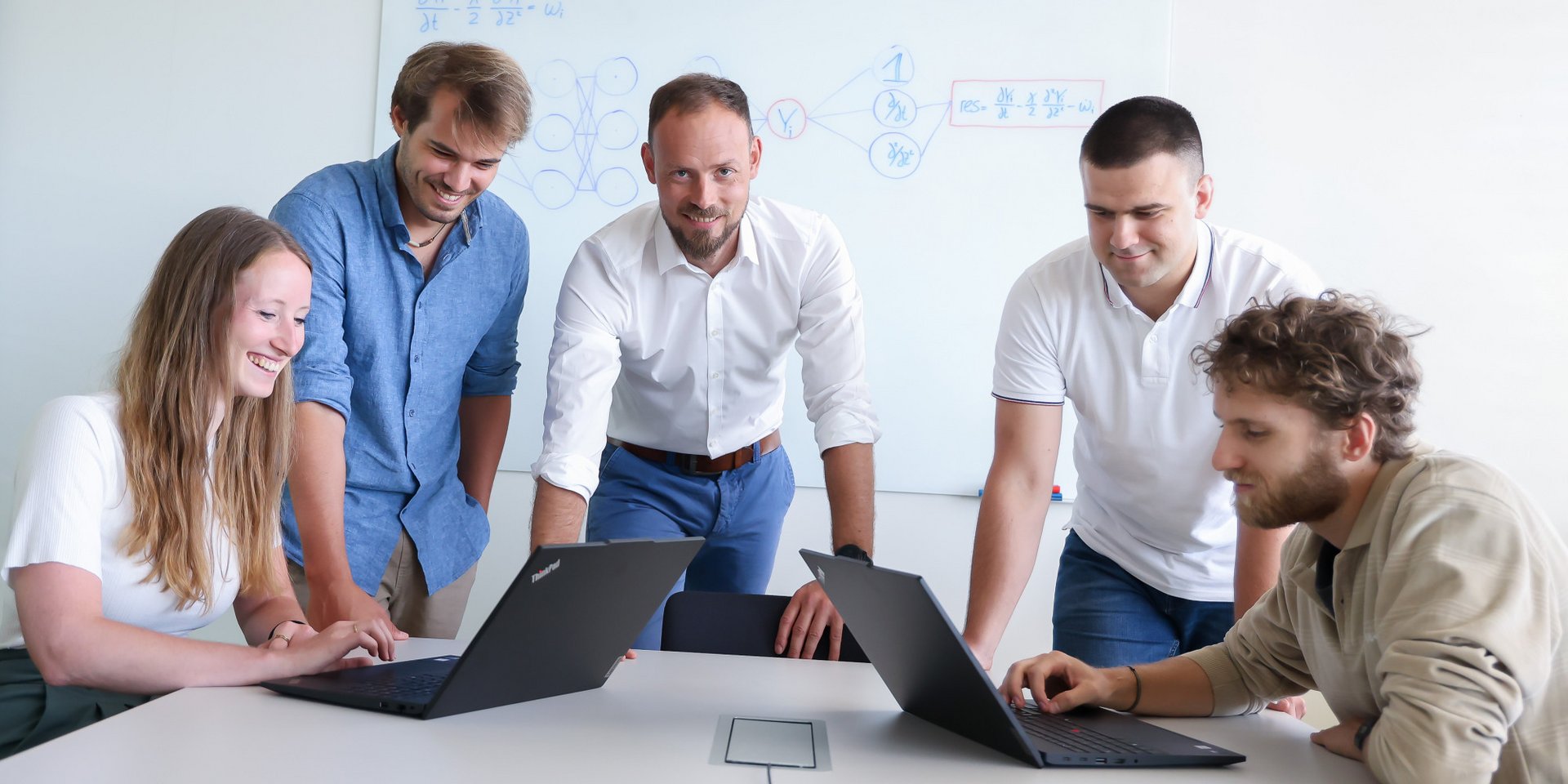 Fünf Menschen befinden sich an einem weißen Tisch. Ganz links sitzt eine Frau an einem Laptop, ganz rechts sitzt ein Mann an einem Laptop. Dazwischen stehen drei Männer. Der Mann in der Mitte blickt in die Kamera, die zwei Personen links blicken auf den linken Laptop, die zwei Personen rechts blicken auf den rechten Laptop.