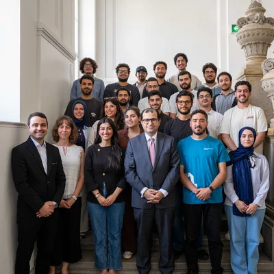 Eine große Gruppe Studierender und Universitätsangehöriger posiert auf einer Stein­treppe mit verzierten Geländern in einem historischen Innenraum. Vorne stehen mehrere Personen in formeller Kleidung, dahinter weitere in legerer Kleidung.