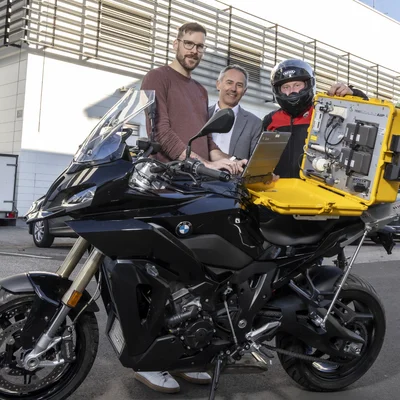 Drei Männer stehen hinter einem Motorrad, auf dem sich ein gelber Koffer mit einem Messgerät befindet.