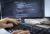 Hände auf einer Computertastatur, im Hintergrund ein Bildschirm mit Code