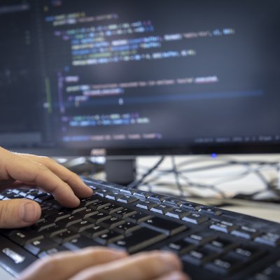Hände auf einer Computertastatur, im Hintergrund ein Bildschirm mit Code