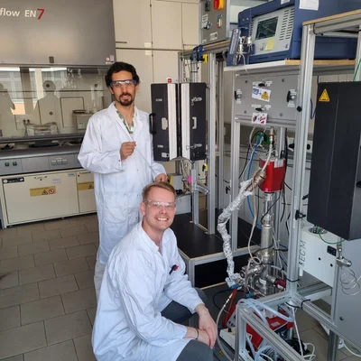 In einem Chemielabor stehen zwei Forschende in Laborkitteln und Schutzbrillen neben einer komplexen Reaktor‑/Messanlage mit Schläuchen, Sensoren und Steuergeräten.