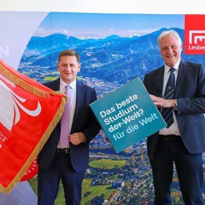 Zwei Männer im Anzug posieren vor einem Panorama‑Hintergrund mit Berg‑/Stadtansicht und Leoben‑Logo. Links wird eine rote Fahne „Österreichischer Städtebund“ gehalten. Rechts halten sie ein Schild mit der Aufschrift „Das beste Studium der Welt? für die Welt“. Gesichter sind unkenntlich gemacht.