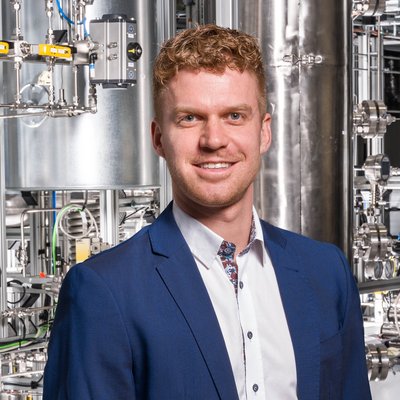 Person in blauem Anzug und weißem Hemd vor einer technischen Anlage mit Edelstahlrohren, Ventilen und Messinstrumenten – industrielles/pilotanlagenartiges Umfeld.