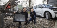 Zwei Männer stehen vor einem Auto und einem Eisenbahnbauteil, der linke Mann hält eine Platte mit Metallkreisen, der rechte Mann einen Verbindungsrüssel