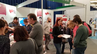 Many - mainly young - people in groups engrossed in conversation in front of display boards at an exhibition stand.