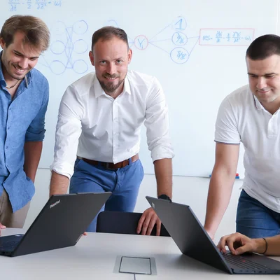 Five people are at a white table. On the far left, a woman is sitting at a laptop; on the far right, a man is sitting at a laptop. Three men are standing between them. The man in the middle is looking at the camera; the two people on the left are looking at the laptop on the left; the two people on the right are looking at the laptop on the right.