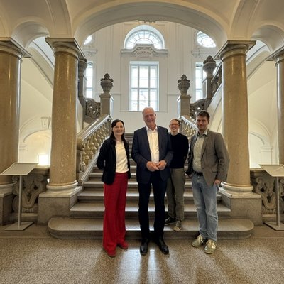 Vier Personen stehen vor einer historischen, säulengerahmten Treppe in einem hellen Foyer; sie blicken in die Kamera.