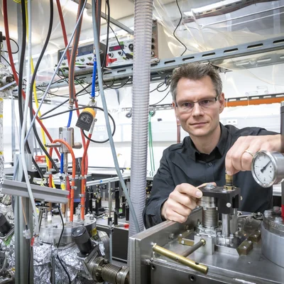 Ein Mann mit Brille steht in einem Labor zwischen mehreren Schläuchen und Metallzylindern.