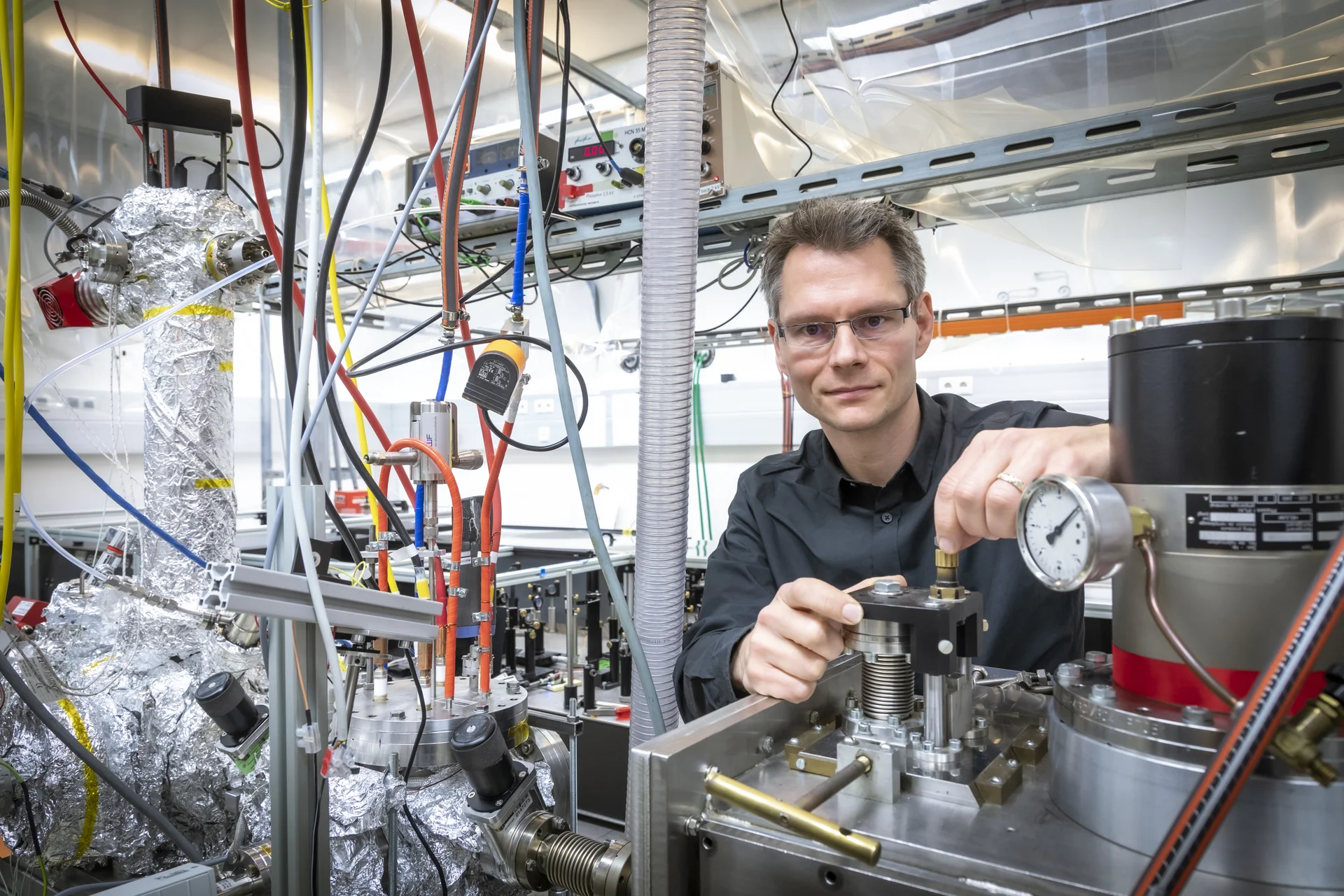 Ein Mann mit Brille steht in einem Labor zwischen mehreren Schläuchen und Metallzylindern.