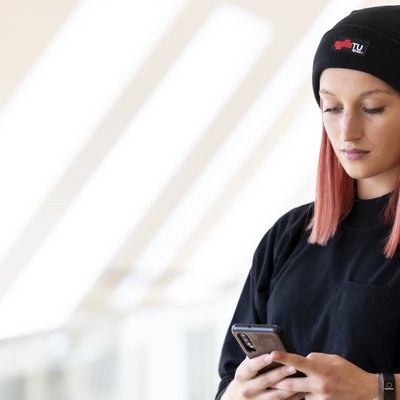 Eine Frau mit einer schwarzen Haube und einem schwarzen T-Shirt blickt konzentriert auf ein Smartphone.