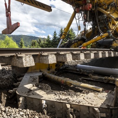 Ausschnitt einer Baumaschine auf Schienen, die ein Stück eines Gleiskörpers anhebt.